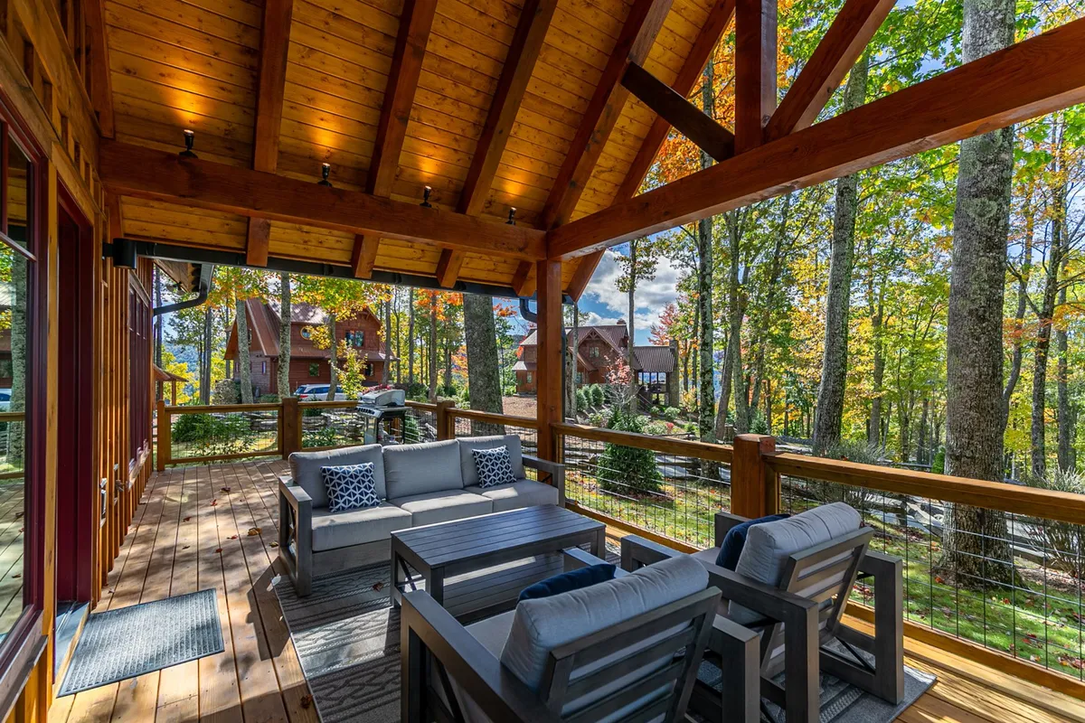 An inviting exterior lounge area featuring sturdy wooden beams, plush seating, and a cozy atmosphere. The surrounding autumn foliage provides a vibrant seasonal backdrop while neighboring homes hint at the community setting. The space is ideal for relaxing outdoors, gathering with family, or enjoying the changing scenery throughout the year.