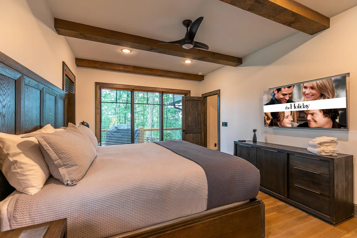 A relaxing king bedroom with exposed wooden beams, a large bed, windows offering a glimpse of nature, and a wall-mounted television for private downtime. The soft, welcoming decor exudes mountain charm.