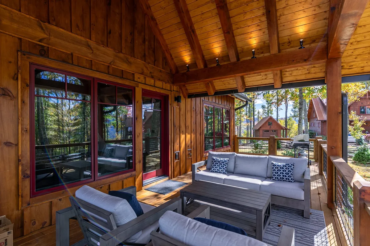 A view of a spacious wooden deck with comfortable seating, adjacent to large windows and surrounded by natural mountain scenery. The deck is set up for relaxed gatherings, providing an ideal spot for outdoor dining or lounging while taking in the expansive views.