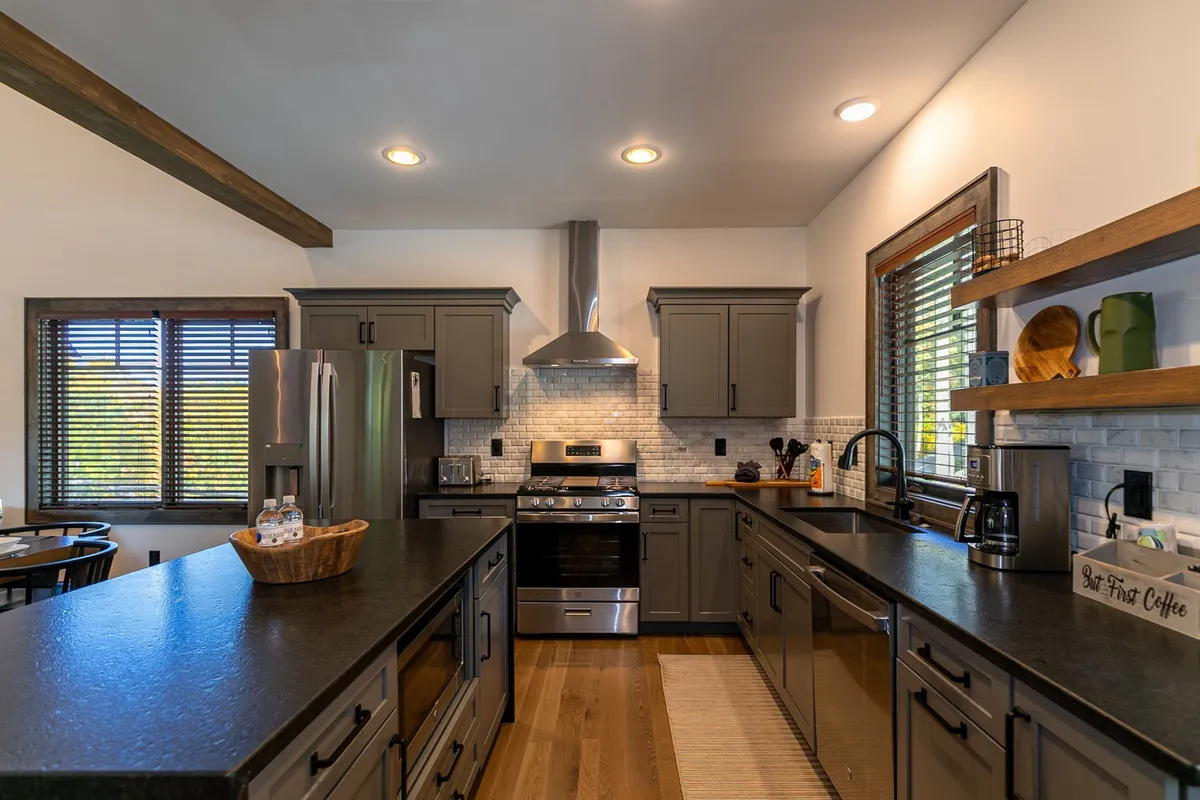 A modern kitchen featuring a prominent island with bar stools, sleek stainless-steel appliances, wooden shelves, and a sunlit window. The setup is ideal for cooking, casual dining, and morning coffee, with a clean, inviting atmosphere.