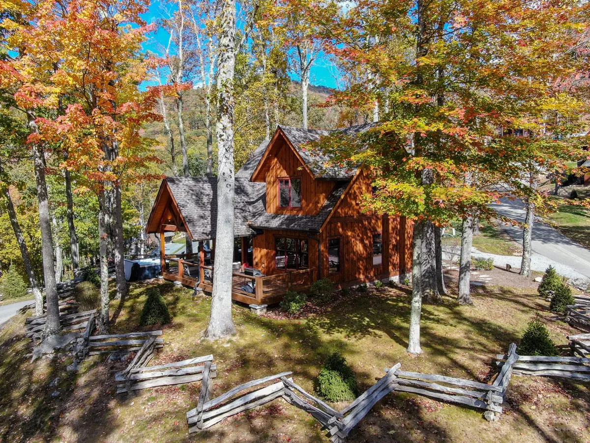 The image showcases a charming wooden cabin nestled among vibrant autumn foliage, with a spacious fenced yard and panoramic views of the surrounding mountains. The inviting porch overlooks a tranquil landscape, perfect for relaxing at sunset or enjoying a cozy evening under the stars.
