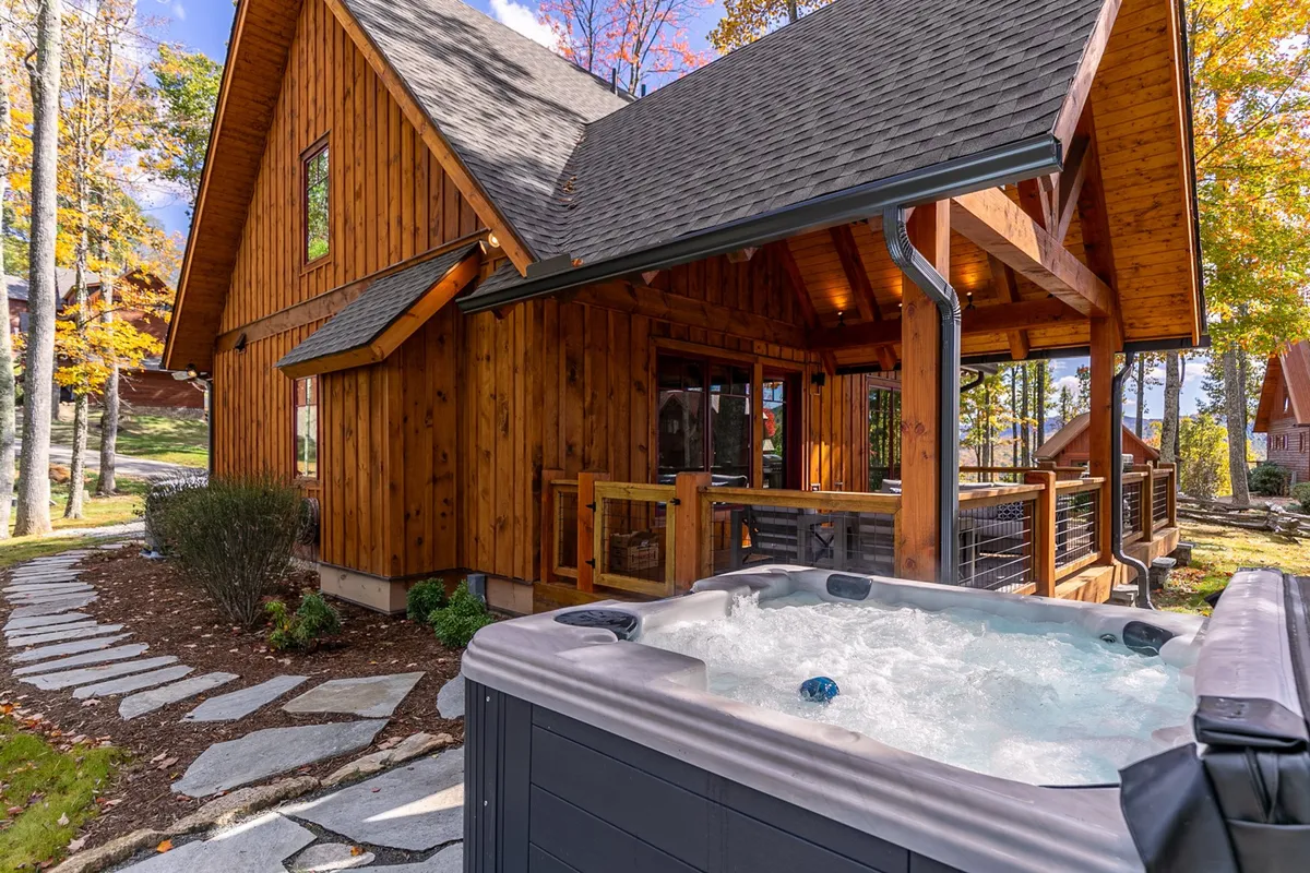 This exterior shot showcases a cozy wooden cabin surrounded by lush trees. A hot tub sits on the deck with a stone pathway leading up, emphasizing both relaxation and connection to the natural environment. The scene feels inviting, with the hot tub placed for optimal views of the landscape and sky, ideal for unwinding after outdoor adventures.