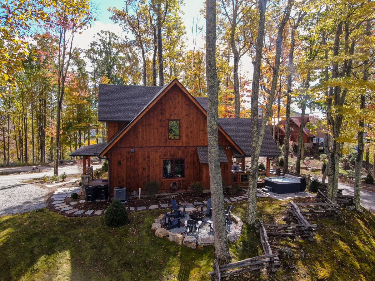 A rustic wooden cabin is surrounded by trees, featuring a deck and a fire pit seating area with a hot tub nearby. The open outdoor amenities and forested backdrop suggest a welcoming, pet-friendly environment where guests can relax after exploring with their dogs.