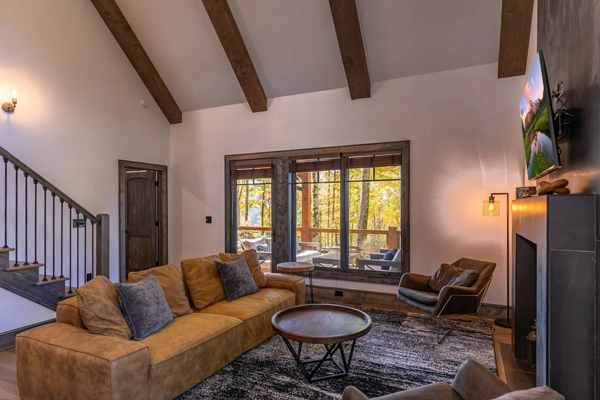 A wide, open living space featuring a large sectional sofa, wooden beams overhead, expansive windows framing breathtaking mountain views, a cozy fireplace, and a swivel chair set in a welcoming layout. The room is styled for comfort and togetherness, perfect for conversation or relaxation.