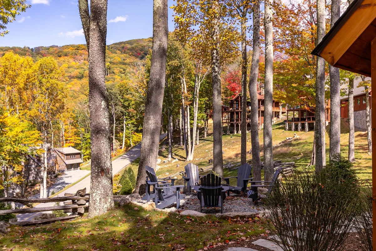A warm, inviting exterior scene featuring Adirondack chairs arranged around a fire pit, set amidst colorful autumn trees and pathways leading to nearby cabins. The setting is ideal for group gatherings by the fire after a day of exploration, with the background showing the broader resort community environment.