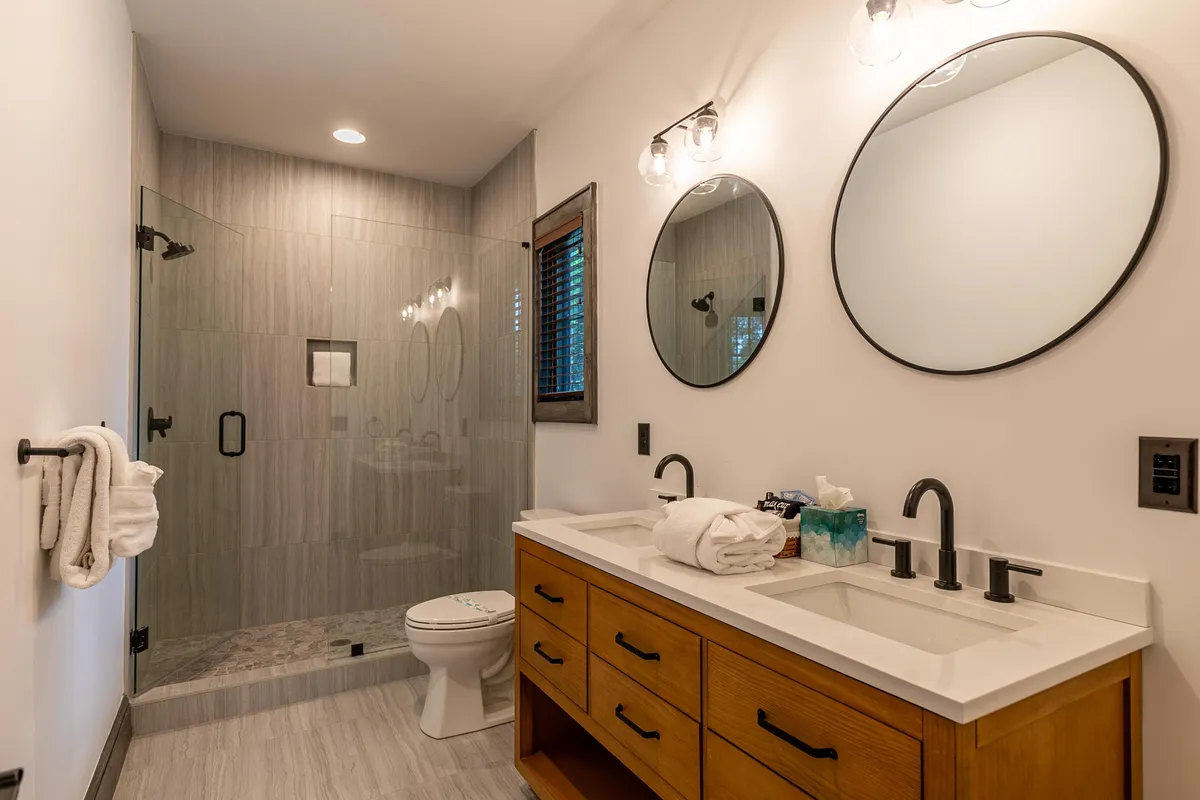 A spa-style bathroom with a double sink vanity, elegant glass shower enclosure, round mirrors, and warm wooden cabinetry. The design is modern, spacious, and tranquil—ideal for a relaxing retreat.