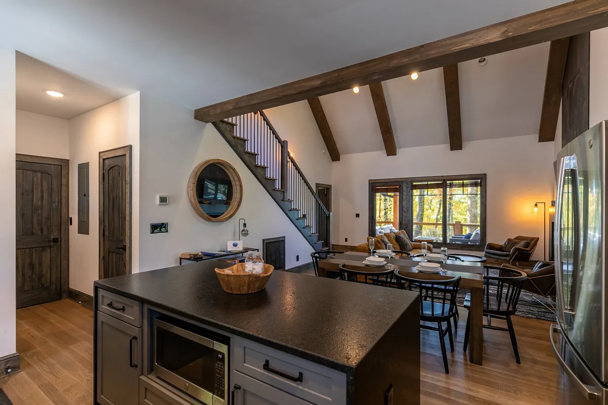 A bright, open space with a kitchen island at the center, adjacent to a dining table, visible staircase, and large windows. The layout highlights the seamless flow between the kitchen, dining, and living areas, creating an inviting setting for socializing, cooking, and enjoying meals with a view.