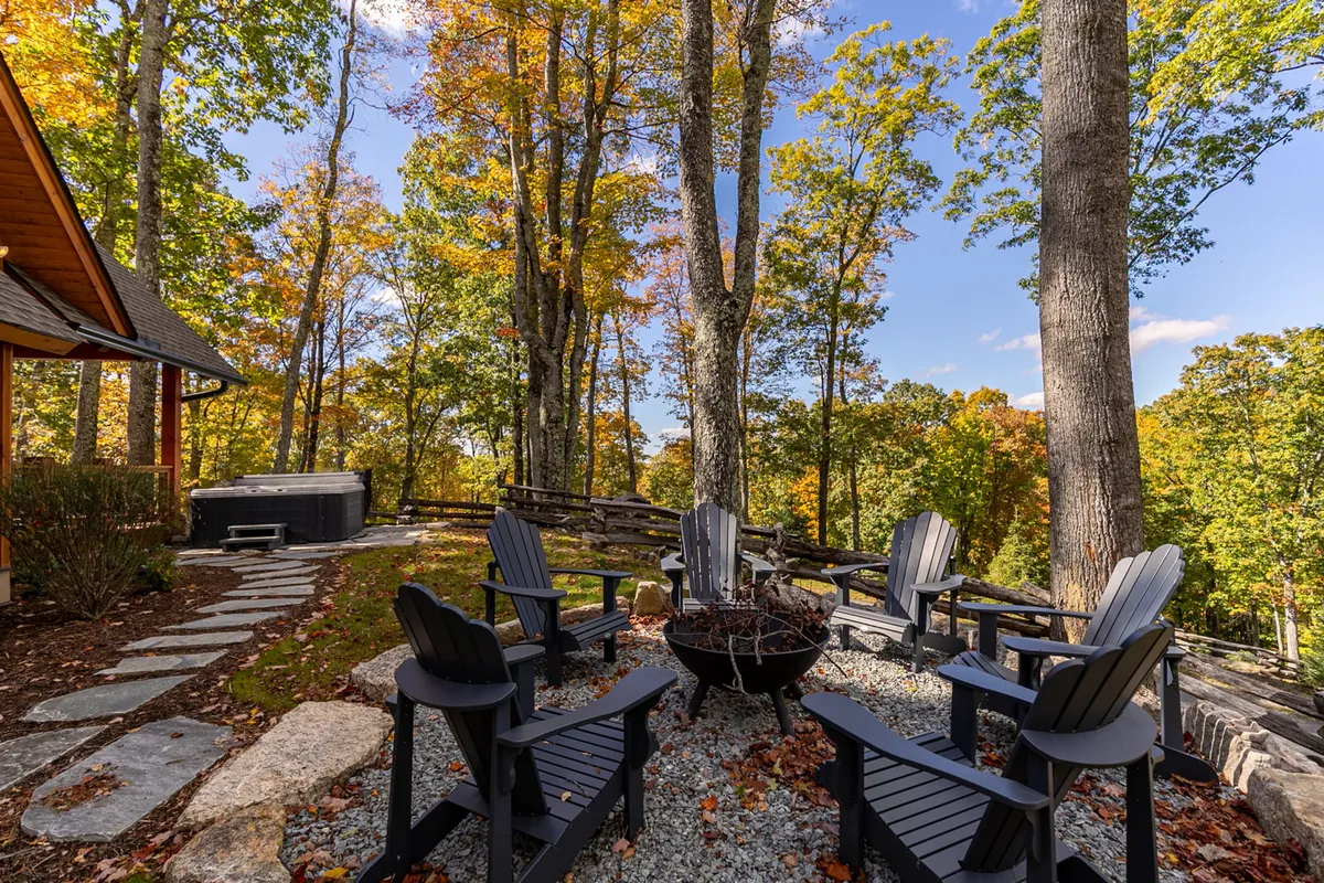 This image features a cozy outdoor space with a fire pit surrounded by Adirondack chairs, and a hot tub tucked nearby. The area is framed by autumn trees, adding a touch of vibrant color and emphasizing the mountain retreat atmosphere. The image clearly presents both the fire pit and hot tub, with ample space for groups to gather, relax, and enjoy the view.